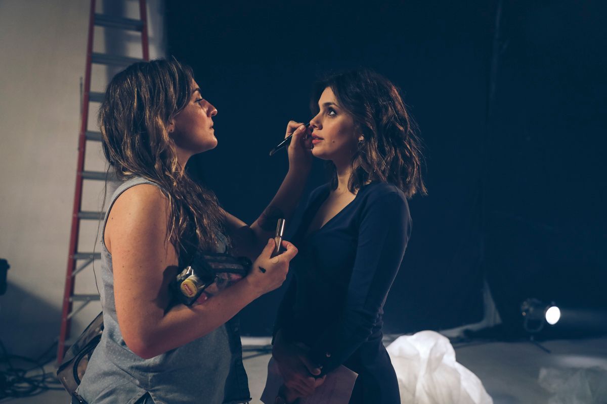 Maquilladora aplicando lápiz labial a una mujer en un estudio. Fondo oscuro, escalera.