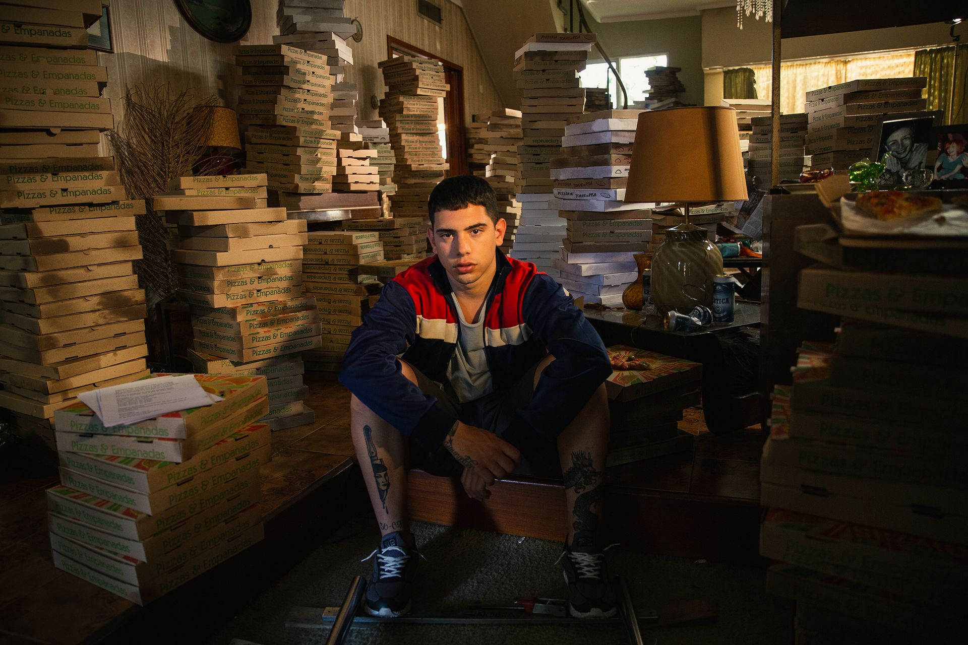 Un joven agachado en una habitación llena de pilas de cajas de pizza, vestido con una chaqueta azul y roja.