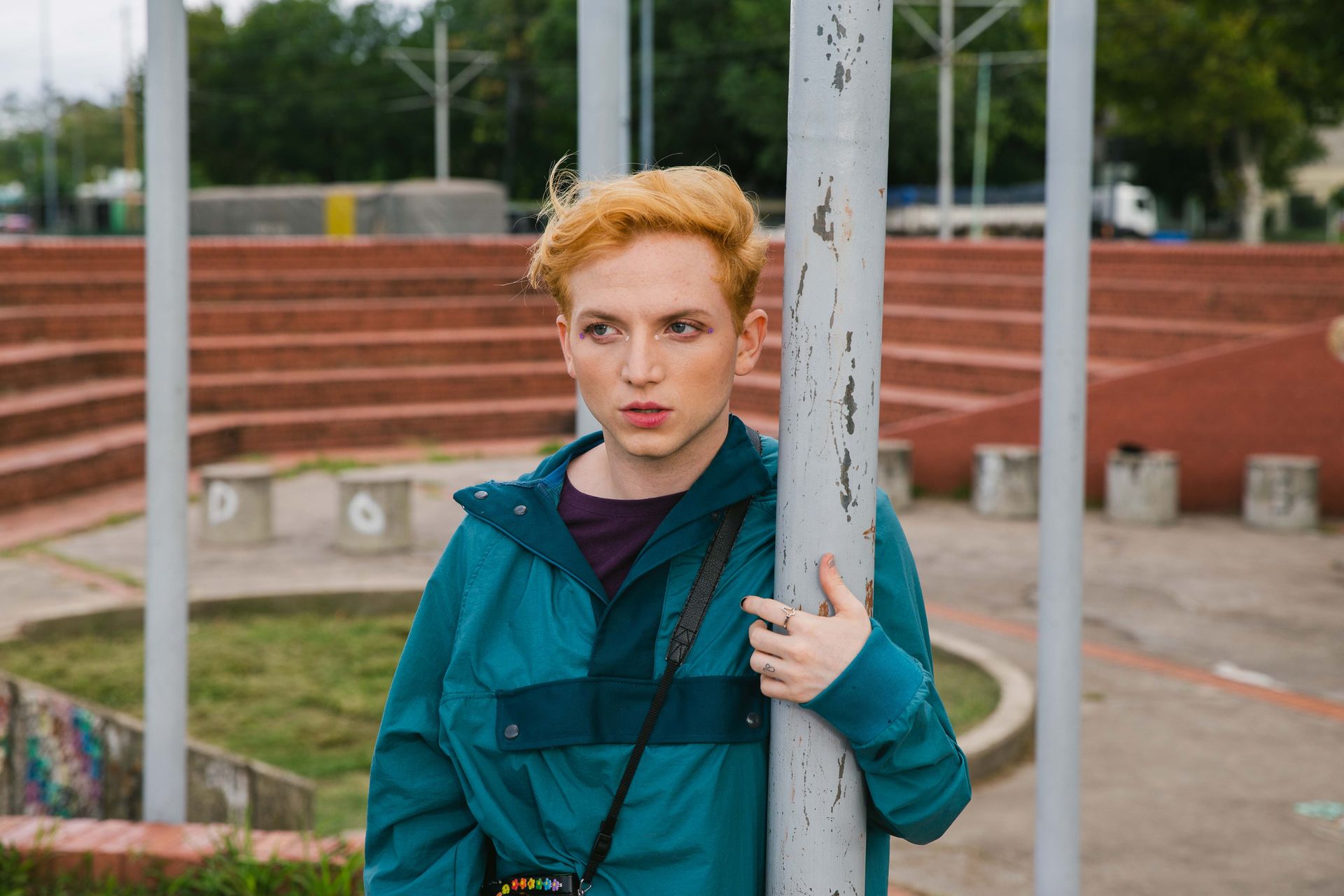 Un joven pelirrojo, apoyado en un poste en un parque público, lleva una chaqueta verde azulada.