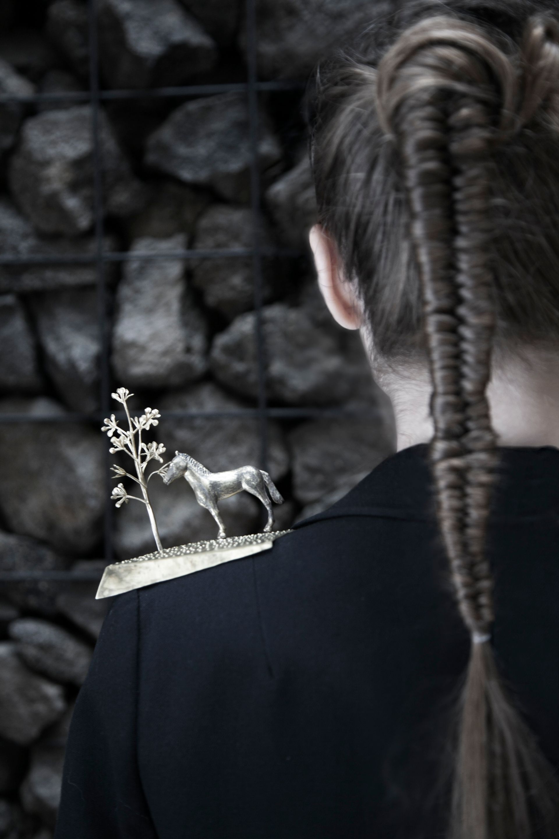 Persona con trenza larga, que lleva un adorno dorado en el hombro de un caballo y un árbol, chaqueta oscura.