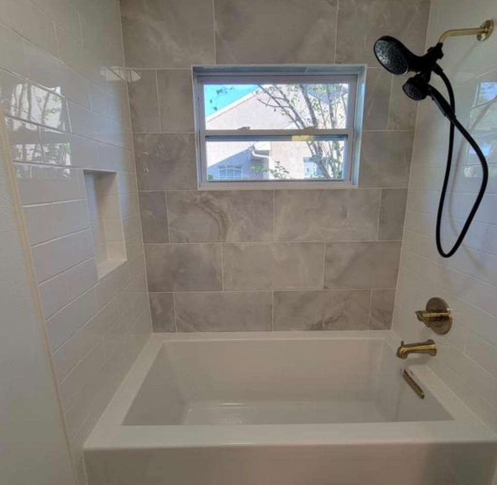 White bathtub and shower with gray tiled accent wall, window, and gold fixtures.