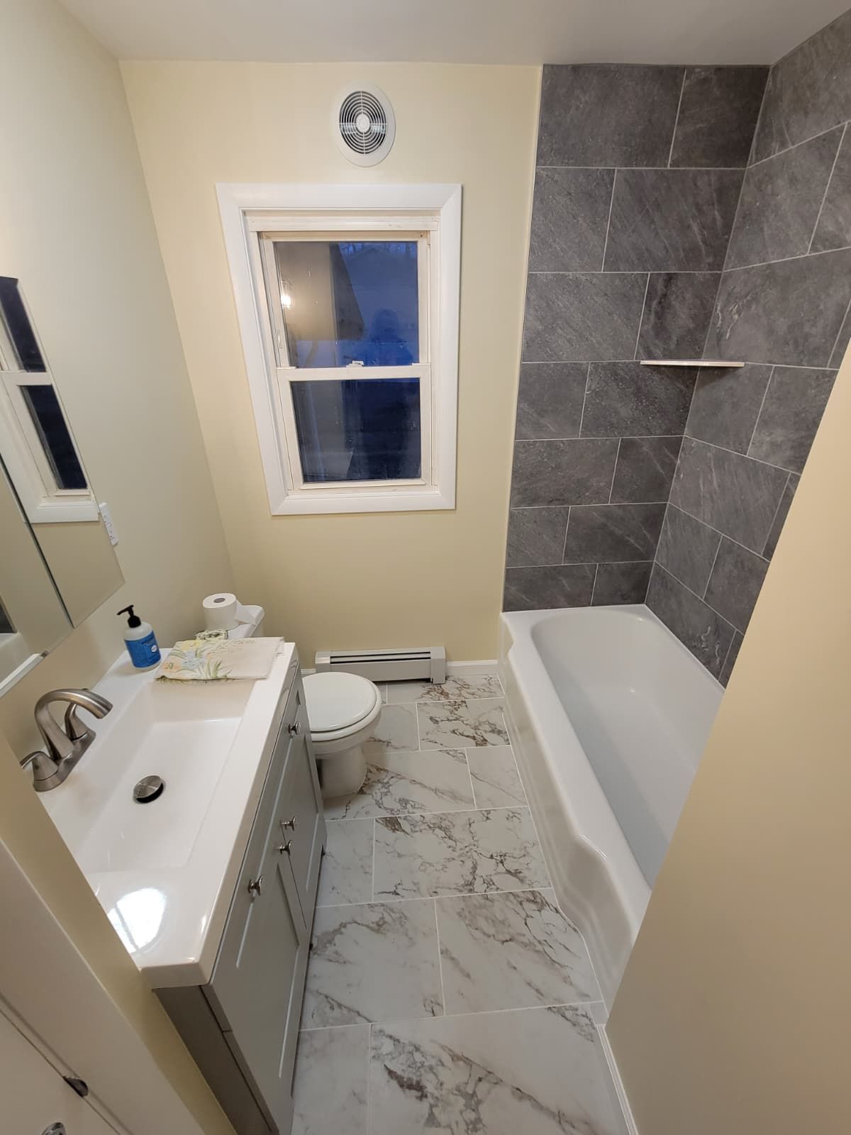 Bathroom with gray tile, white vanity, and tub, window with view of outside.