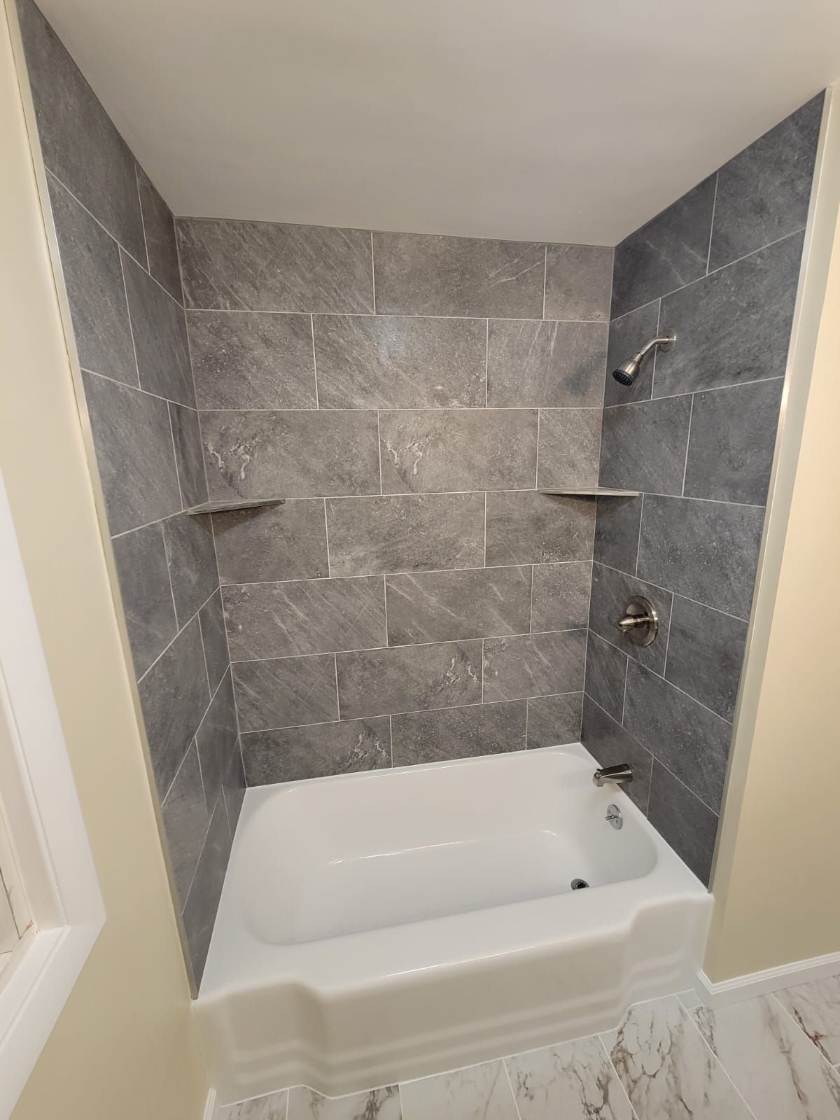 Gray tiled shower with a white bathtub and built-in shelves.