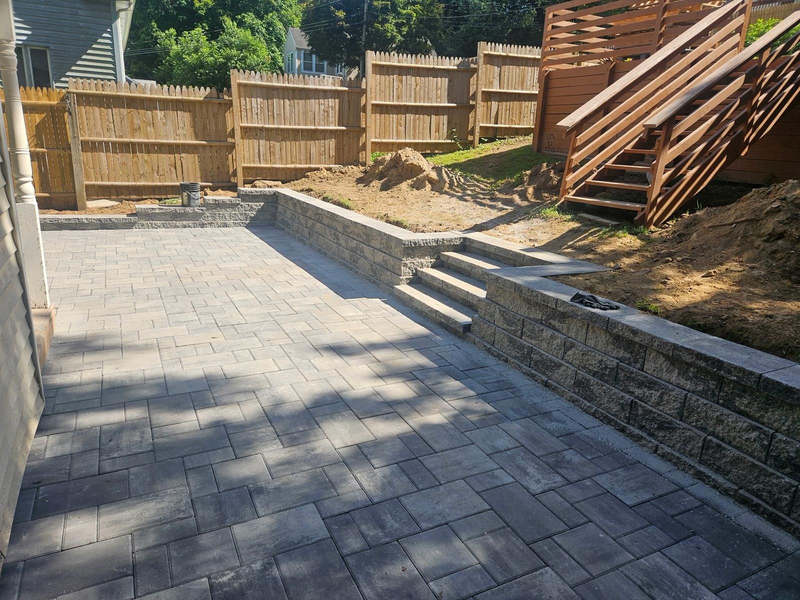 Paver patio and retaining wall with steps, leading to wooden stairs and a wooden fence.