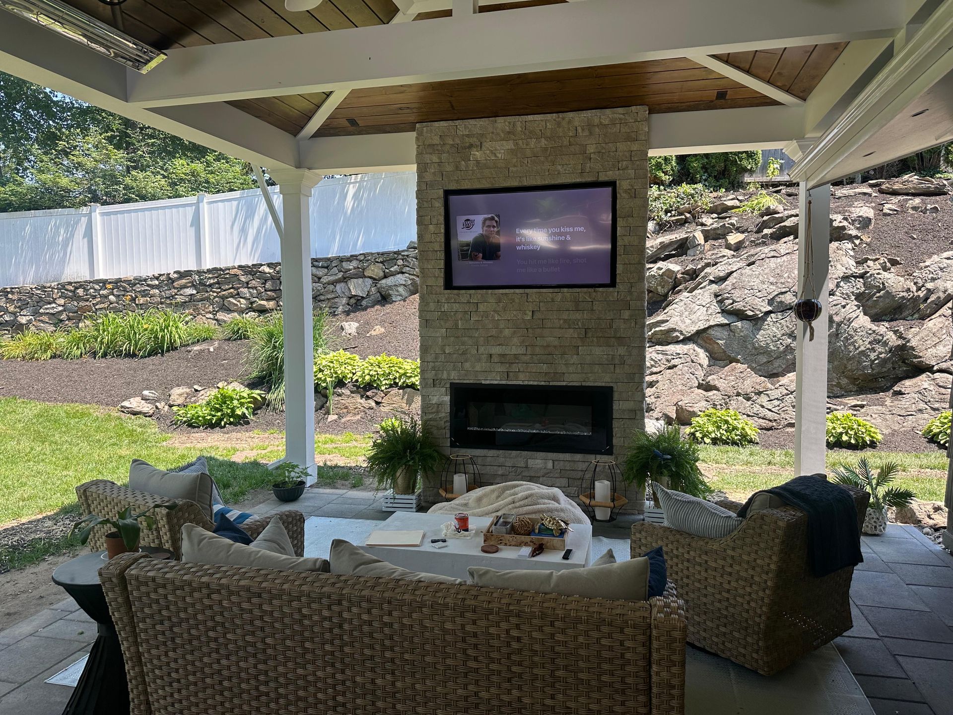 Outdoor living space with stone fireplace, TV, and wicker furniture under a white pergola.
