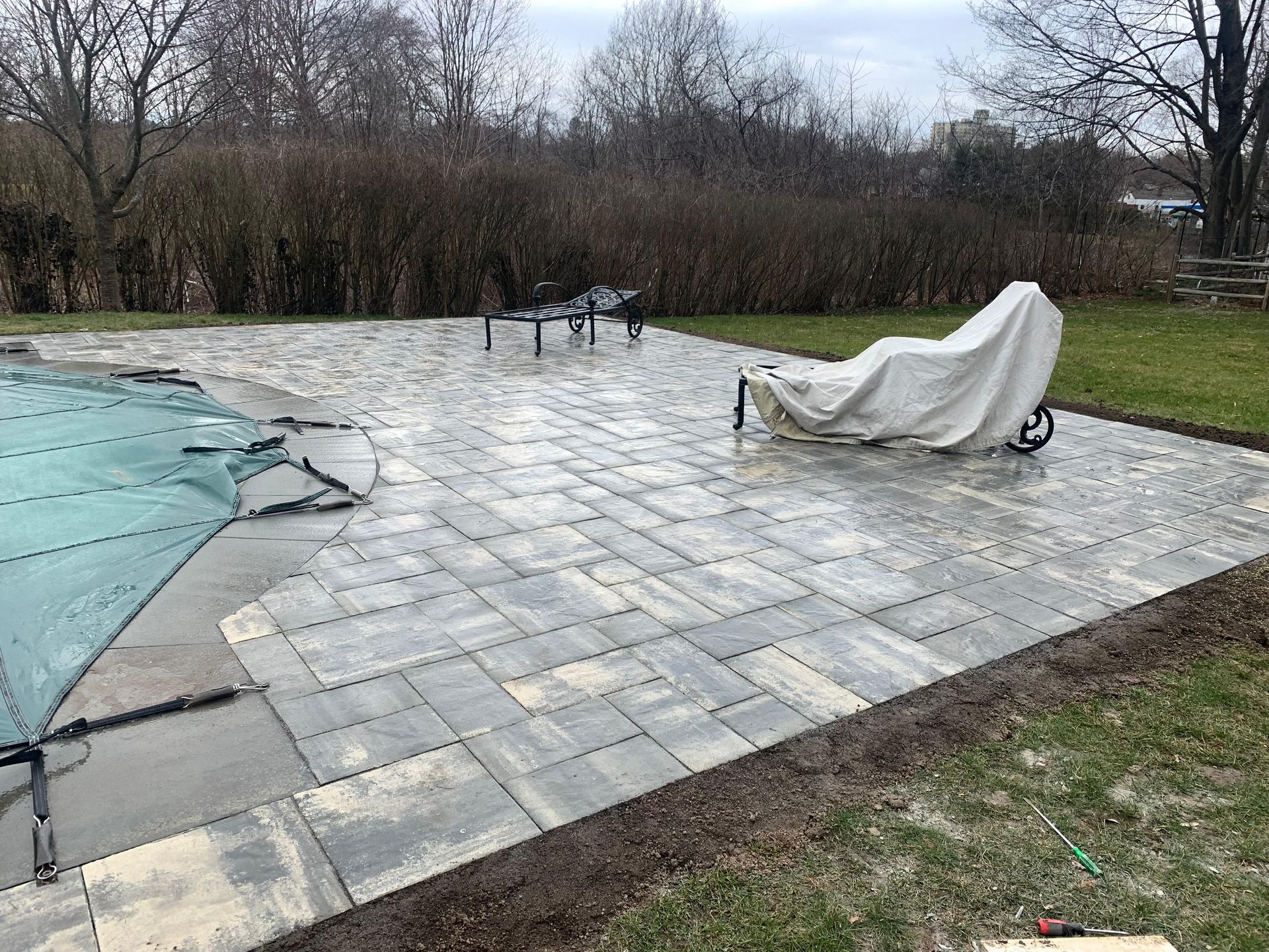 Completed patio with pavers, near a pool, with covered furniture on a cloudy day.