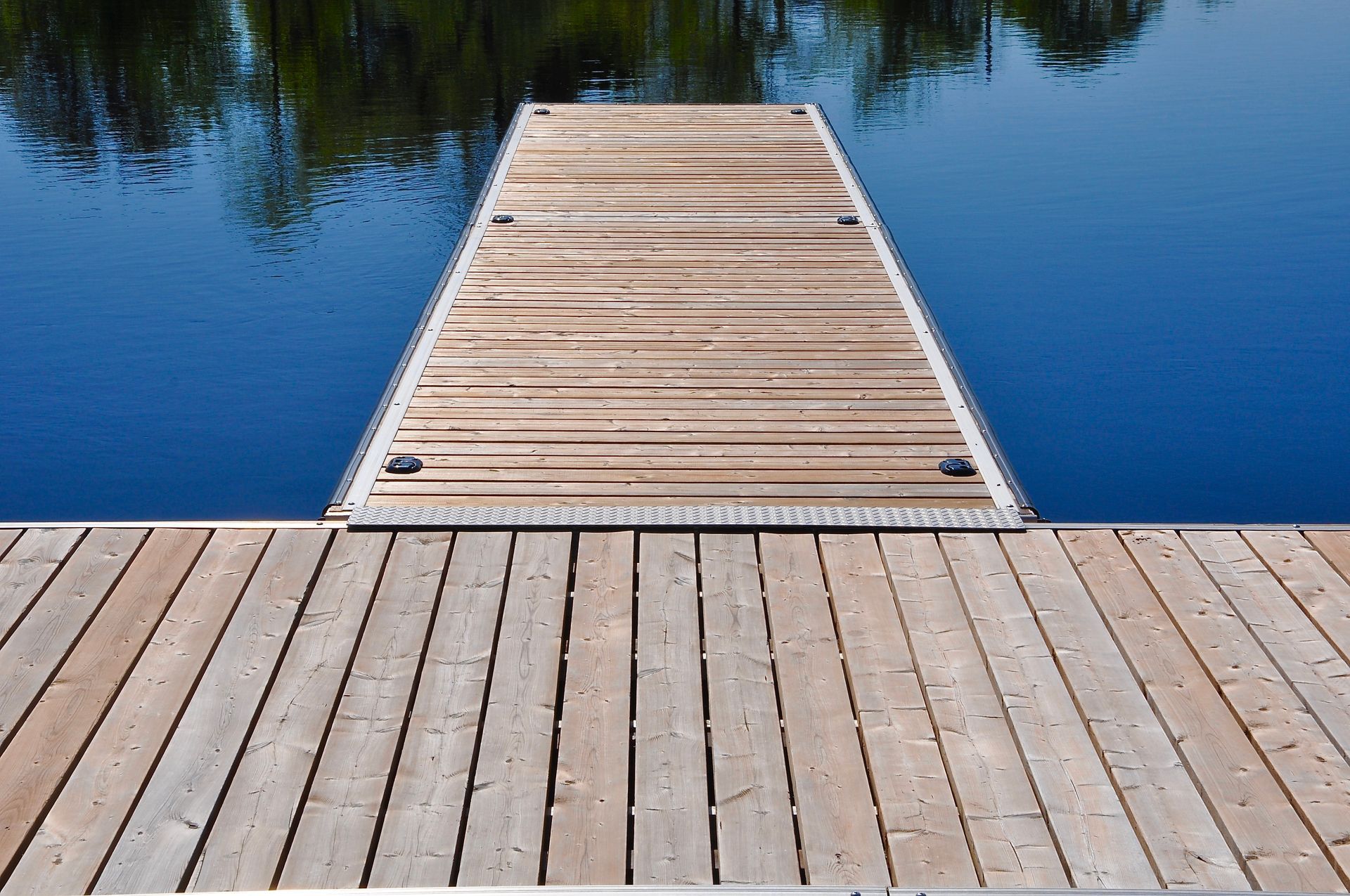 Wooden dock extending into a calm blue lake with tree reflections and a sandy shoreline in the foreground