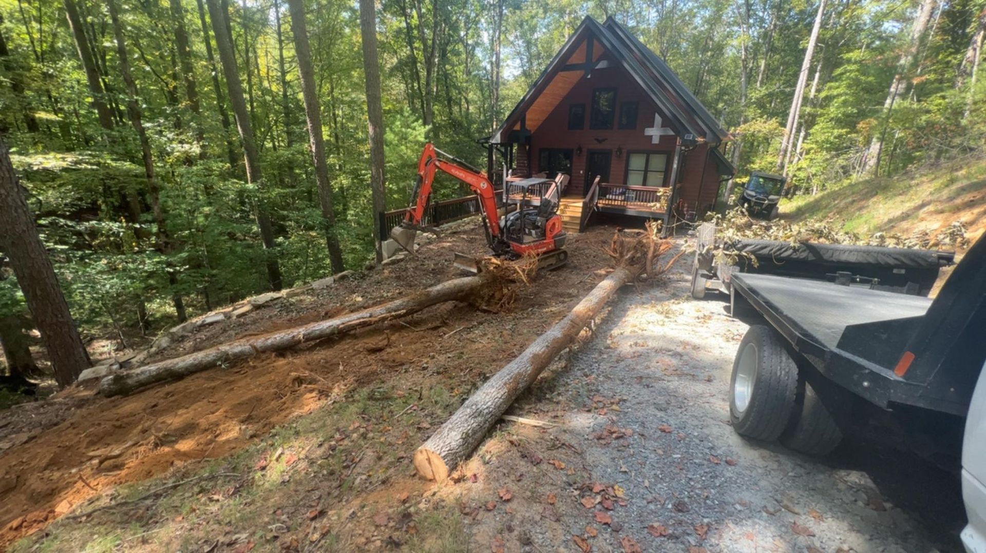 Mini excavator clearing a dirt path lined with logs, beside a wood cabin in a forest.
