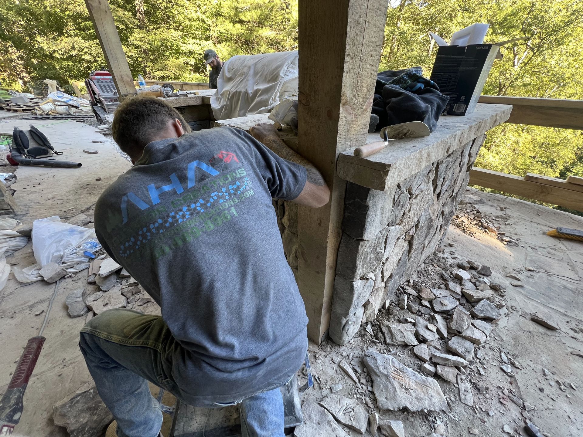 A man in gray shirt kneels, working on a stone structure outdoors. Sunlight and rubble are visible.