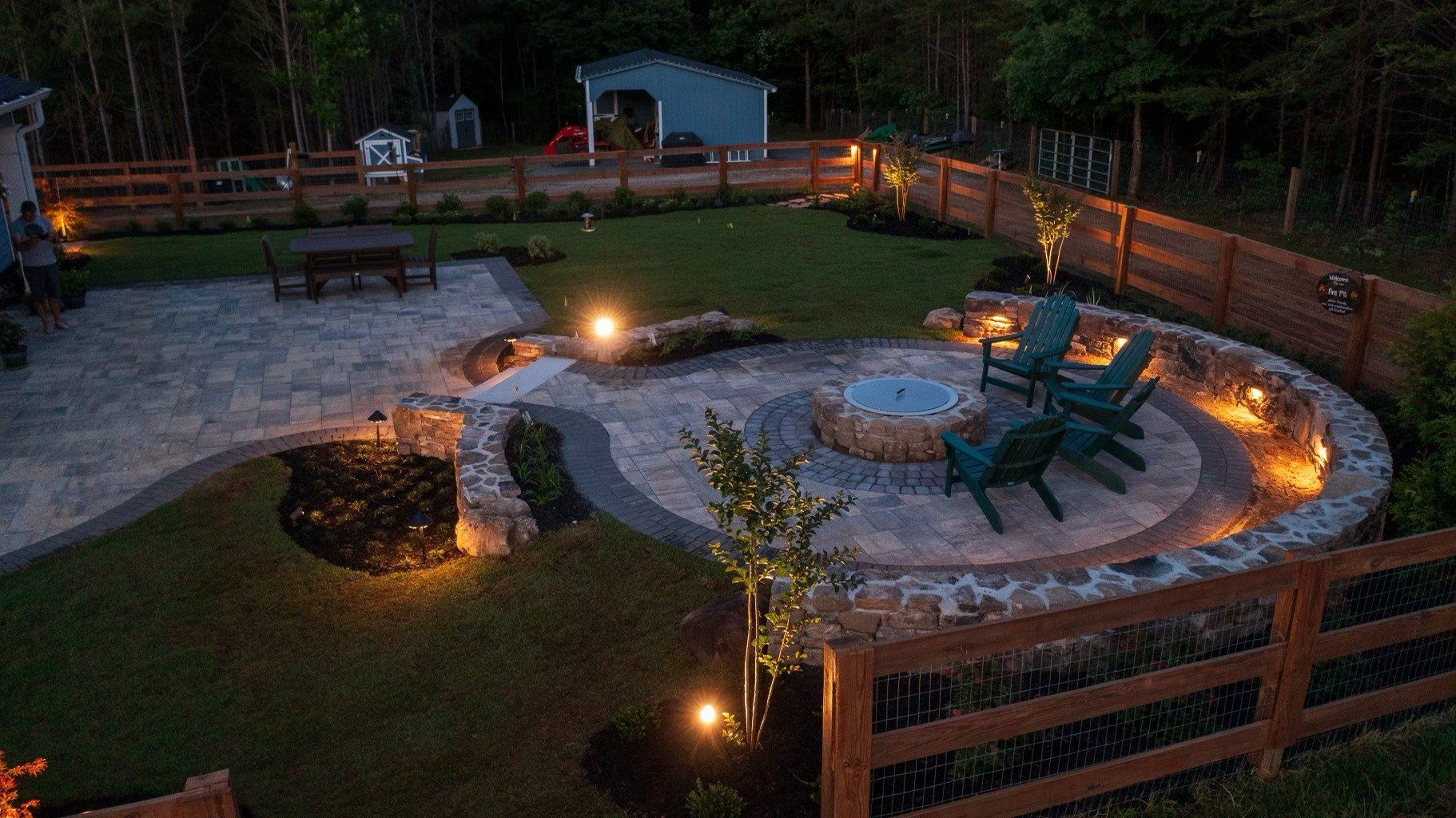 Nighttime outdoor patio with fire pit, stone walls, Adirondack chairs, and pathway lighting.