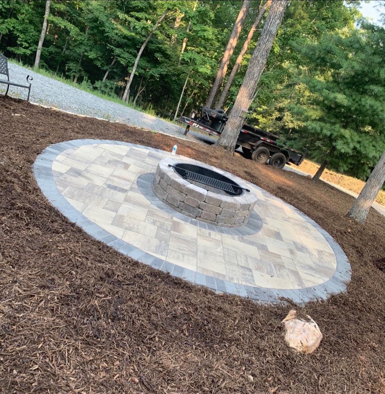 A fire pit is in the middle of a circular patio surrounded by mulch and trees.