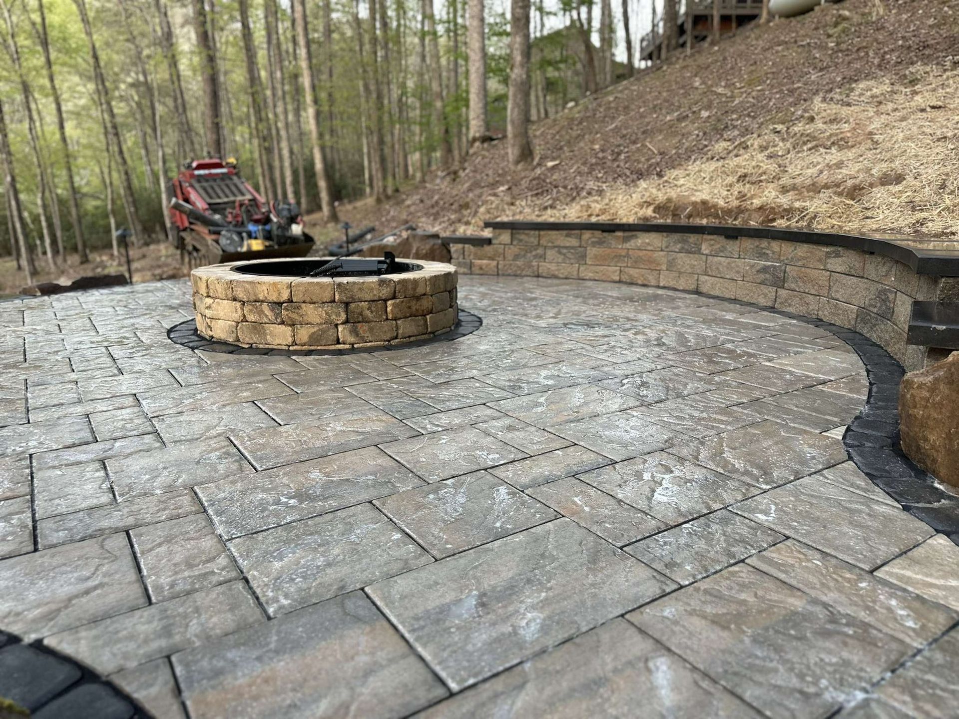 Stone patio with fire pit, retaining wall, and mini excavator in a wooded area.