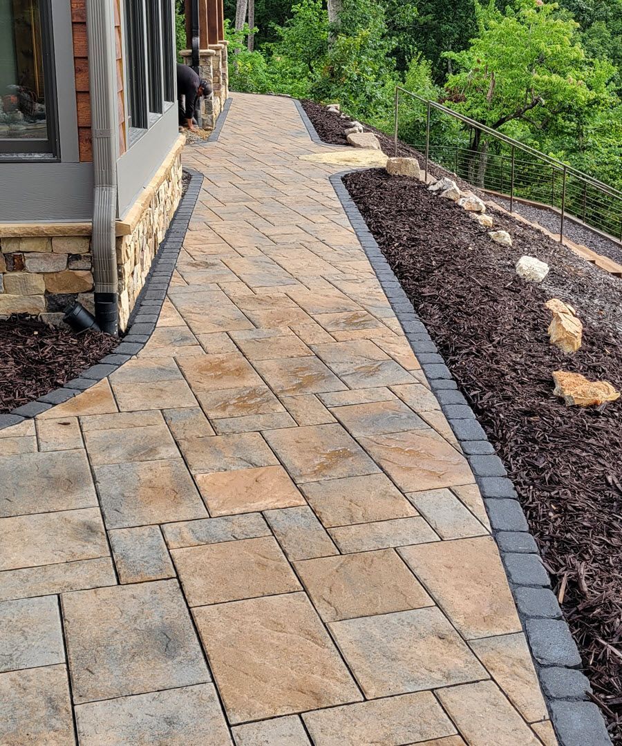 Brick walkway alongside a building, bordered by dark pavers and mulch, leading up a hill.