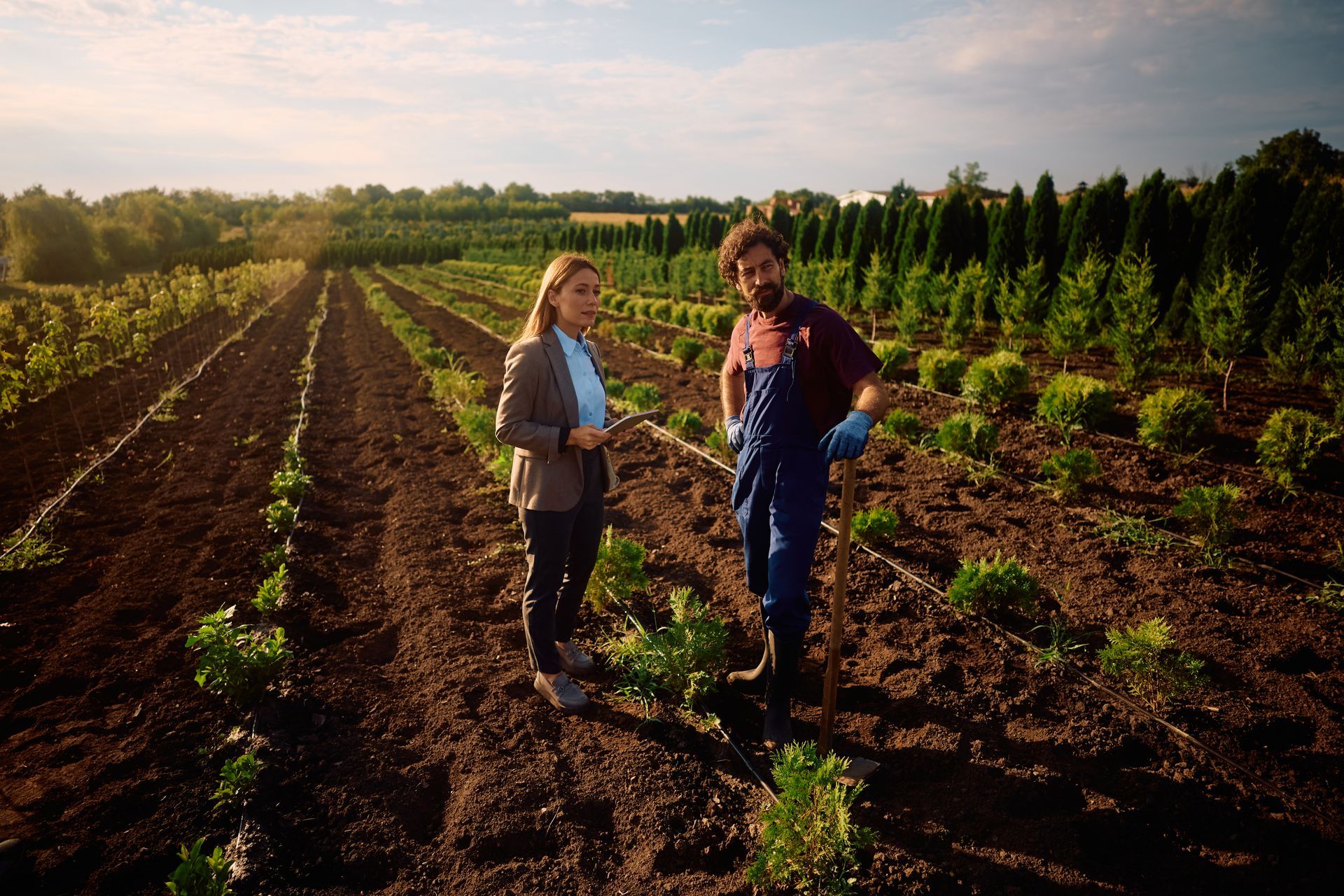 Woman and farmer standing in a field of young plants. The farmer wears overalls; the woman a jacket. Trees in the background.