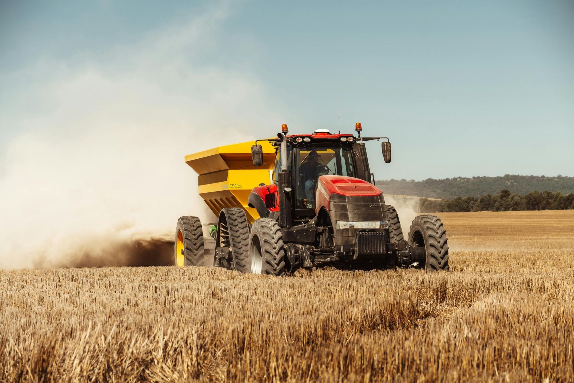 Red tractor with yellow spreader fertilizing a wheat field under a blue sky.