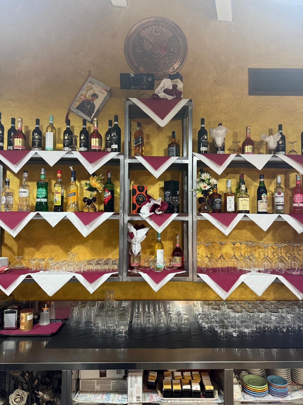 Bar con parete dorata, scaffali pieni di bottiglie di liquore, tovaglioli rossi piegati e bancone.