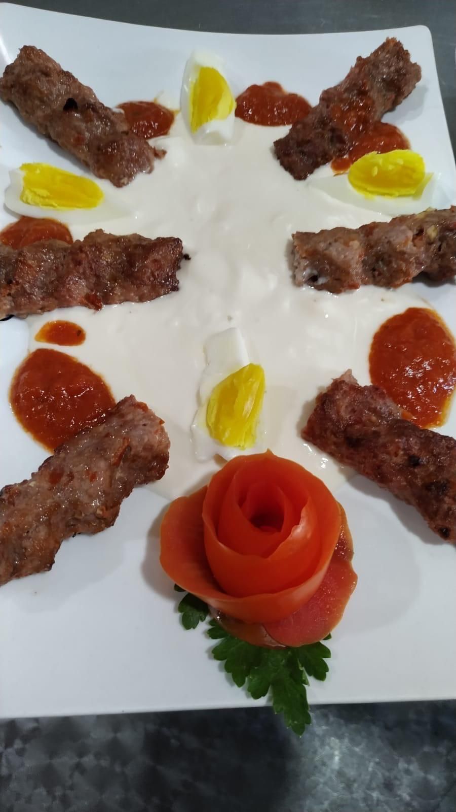 Un piatto con kebab, salsa, metà uova e una rosa di pomodoro, disposti su una salsa bianca.