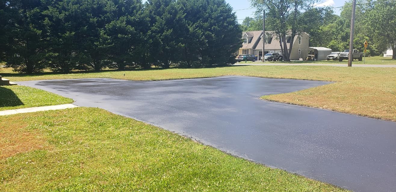 Driveway freshly sealed with lush green grass and trees surrounding. A house sits in the background.