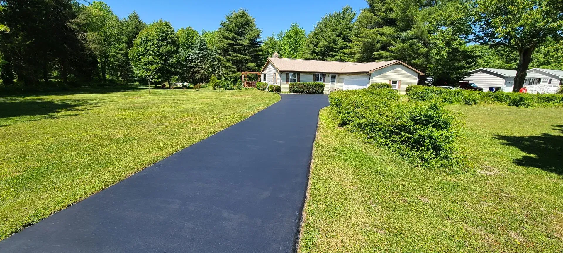 Asphalt driveway leads to a house with a grassy lawn and trees in the background.