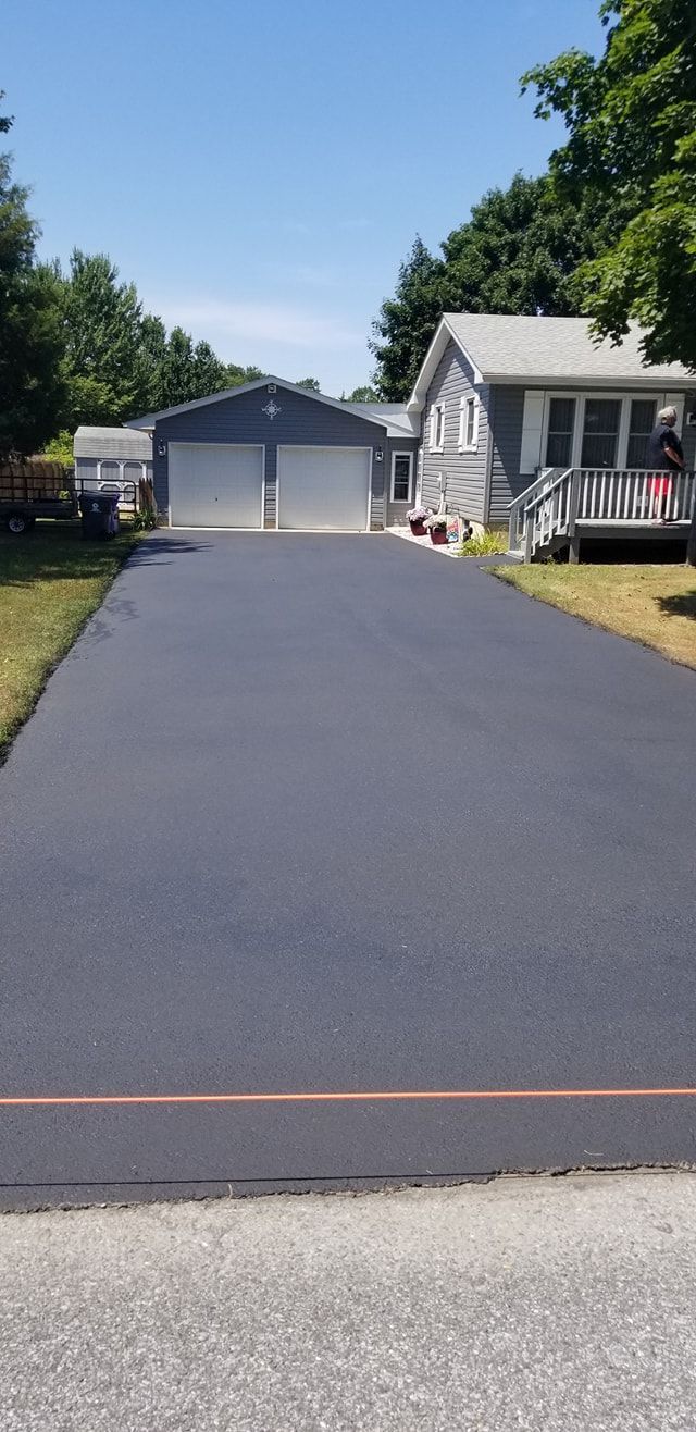 Freshly paved driveway leads to a house and garage. Blue sky, green trees, and a sunny day.