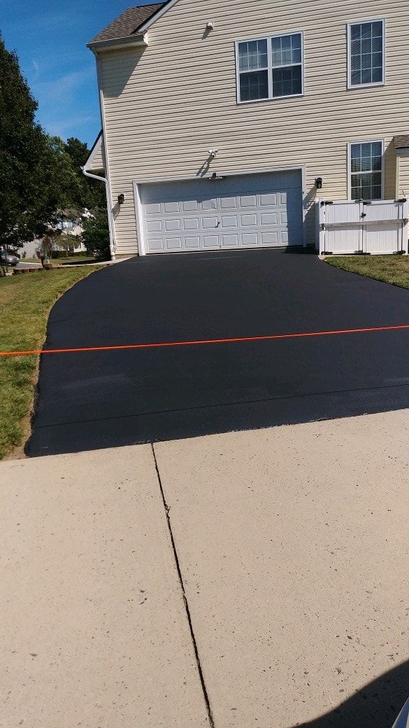 Black asphalt driveway leads to a white garage door on a beige-sided house; a red barrier tape divides the driveway.