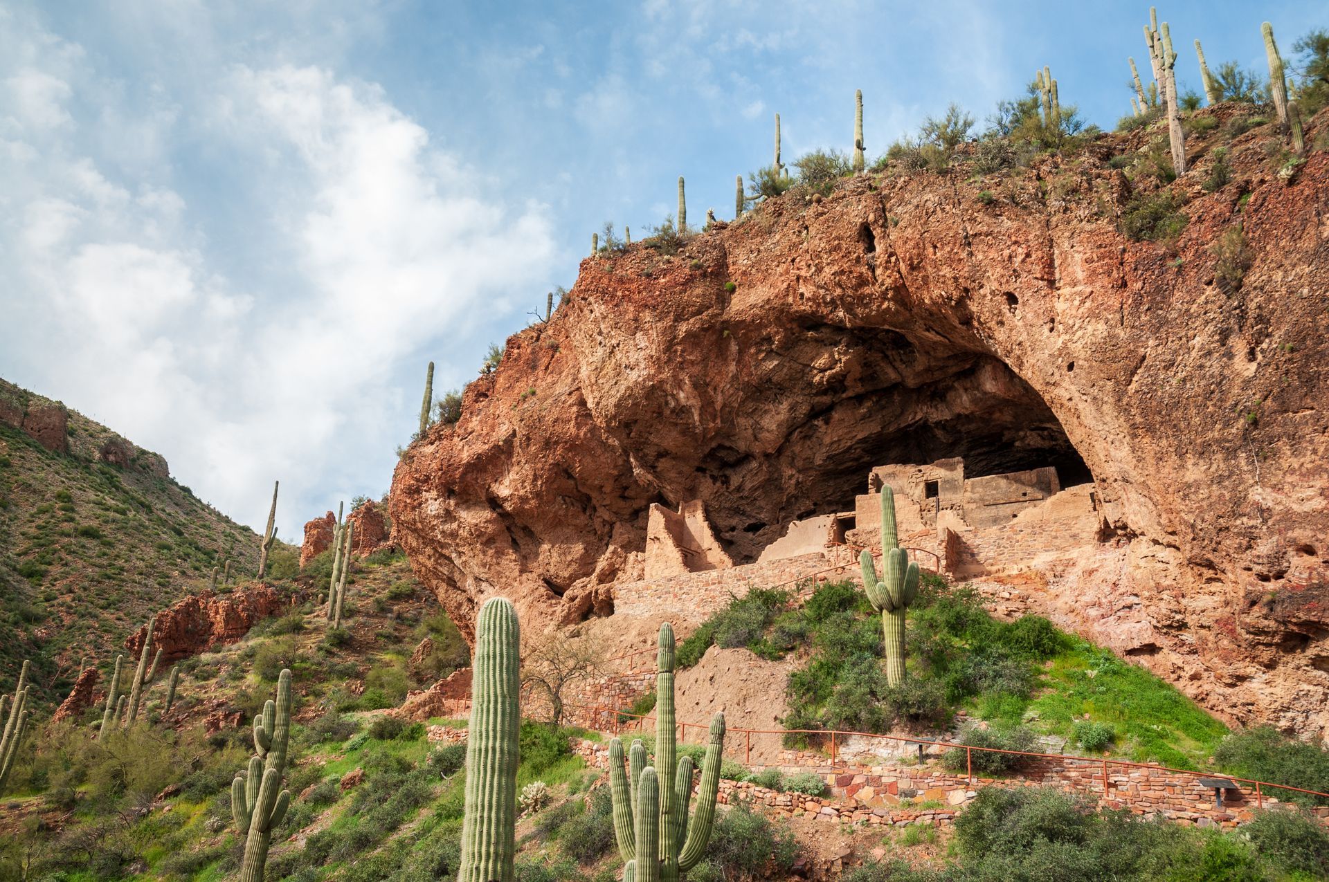 Red rock cave in a desert hillside with cacti and green shrubs under a blue sky