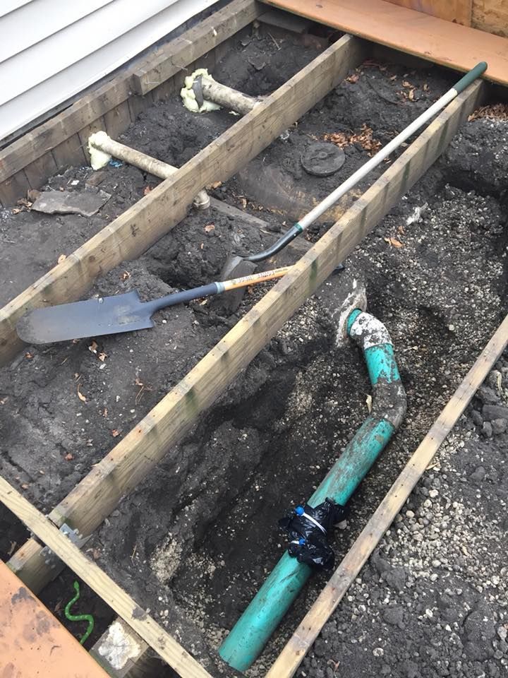 A deck's wooden frame, earth, and plumbing visible. A shovel and other tools lie within the dug-out earth.