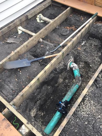 A deck's wooden frame, earth, and plumbing visible. A shovel and other tools lie within the dug-out earth.