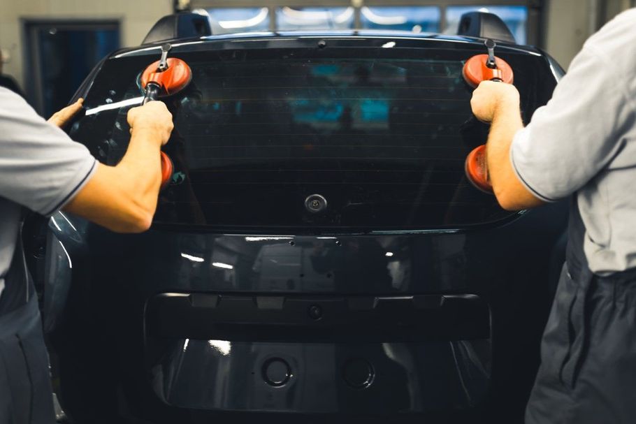 Two workers in gloves polish a black car’s rear with orange buffers in a garage.
