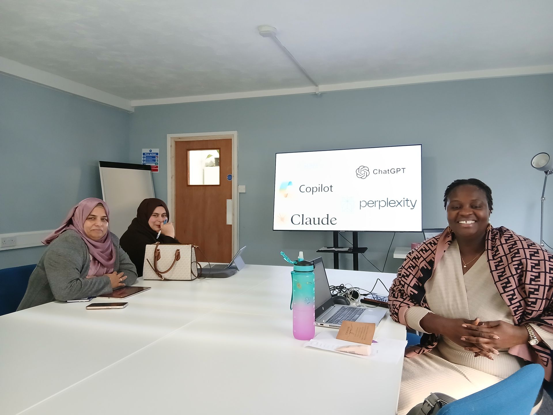 Three women are sitting around a table. They each have a tablet or laptop in front of them. There is a screen with examples of AI software such as 'Chat GPT' and 'Copilot'. 