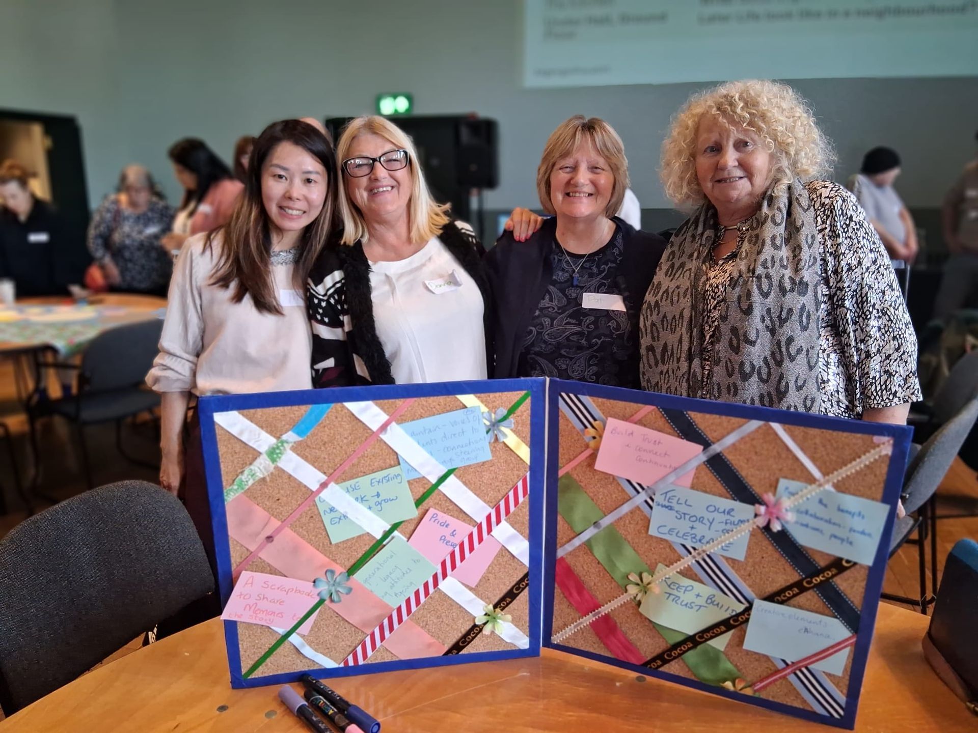 4 older women standing behind a noticeboard covered in handwritten. They are arm in arm and smiling.