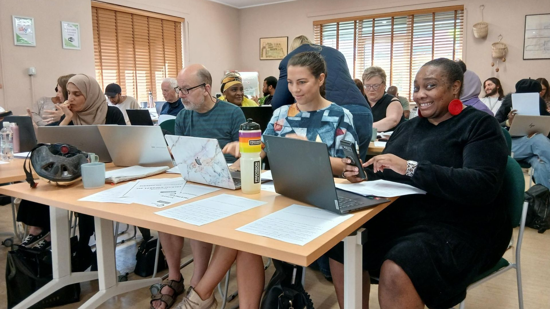 A room full of people sit at desks with a mobile device each, and take part in a workshop.