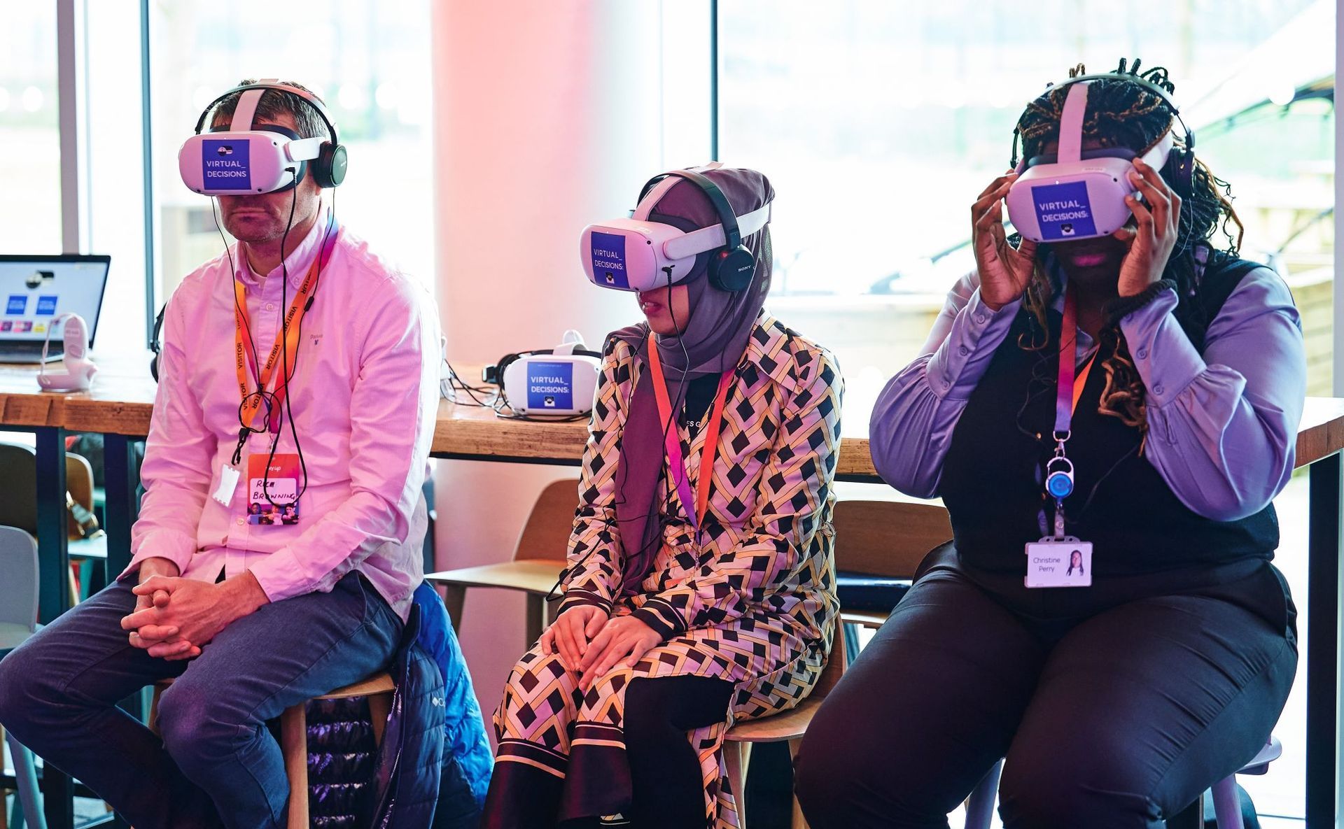 A man and two women sit on stools. They are wearing virtual reality headsets. 
