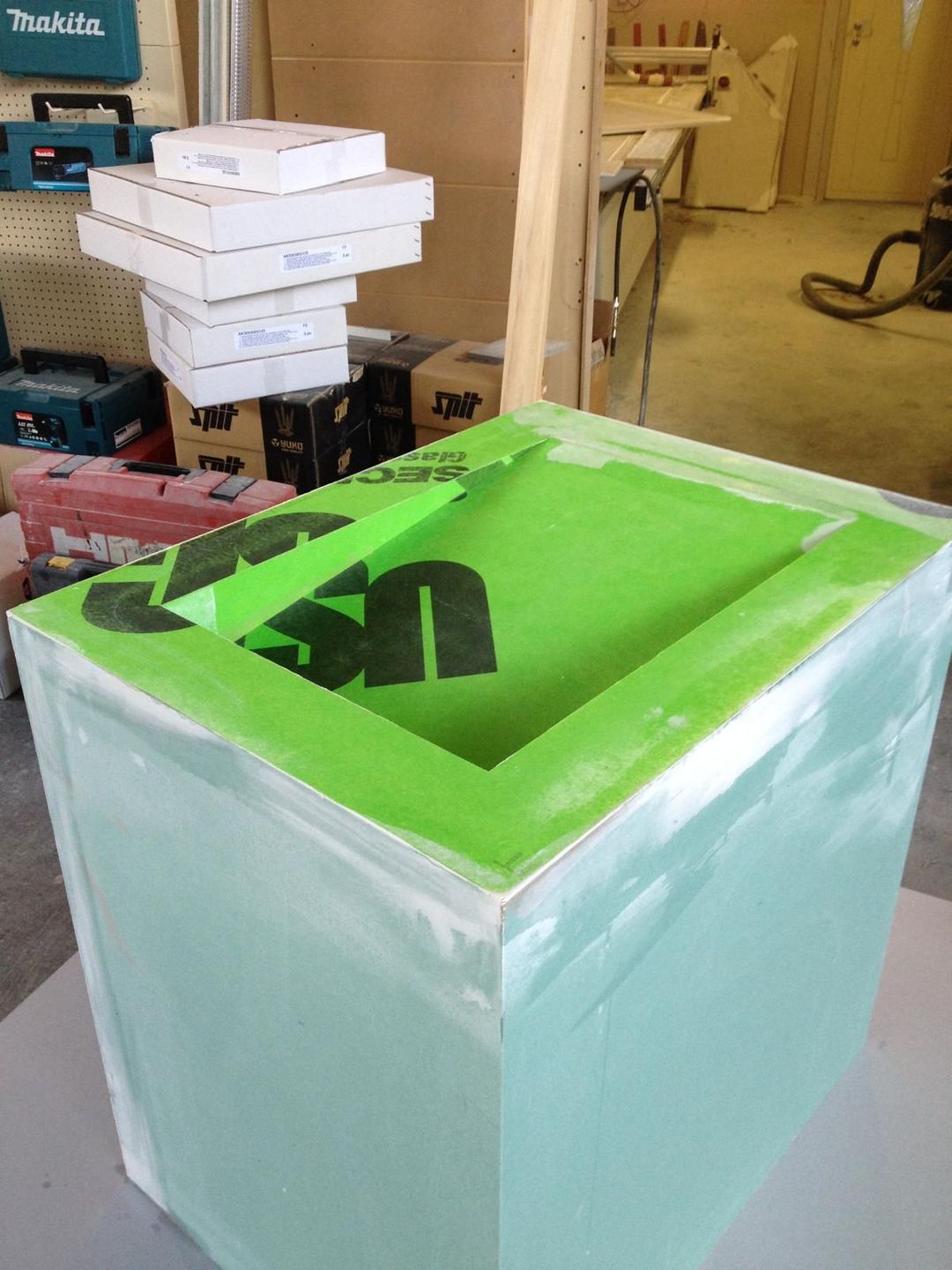 Una struttura a forma di armadio in schiuma verde, sormontata da una cornice quadrata fissata con nastro adesivo, collocata all'interno di un'officina con scatole sullo sfondo.