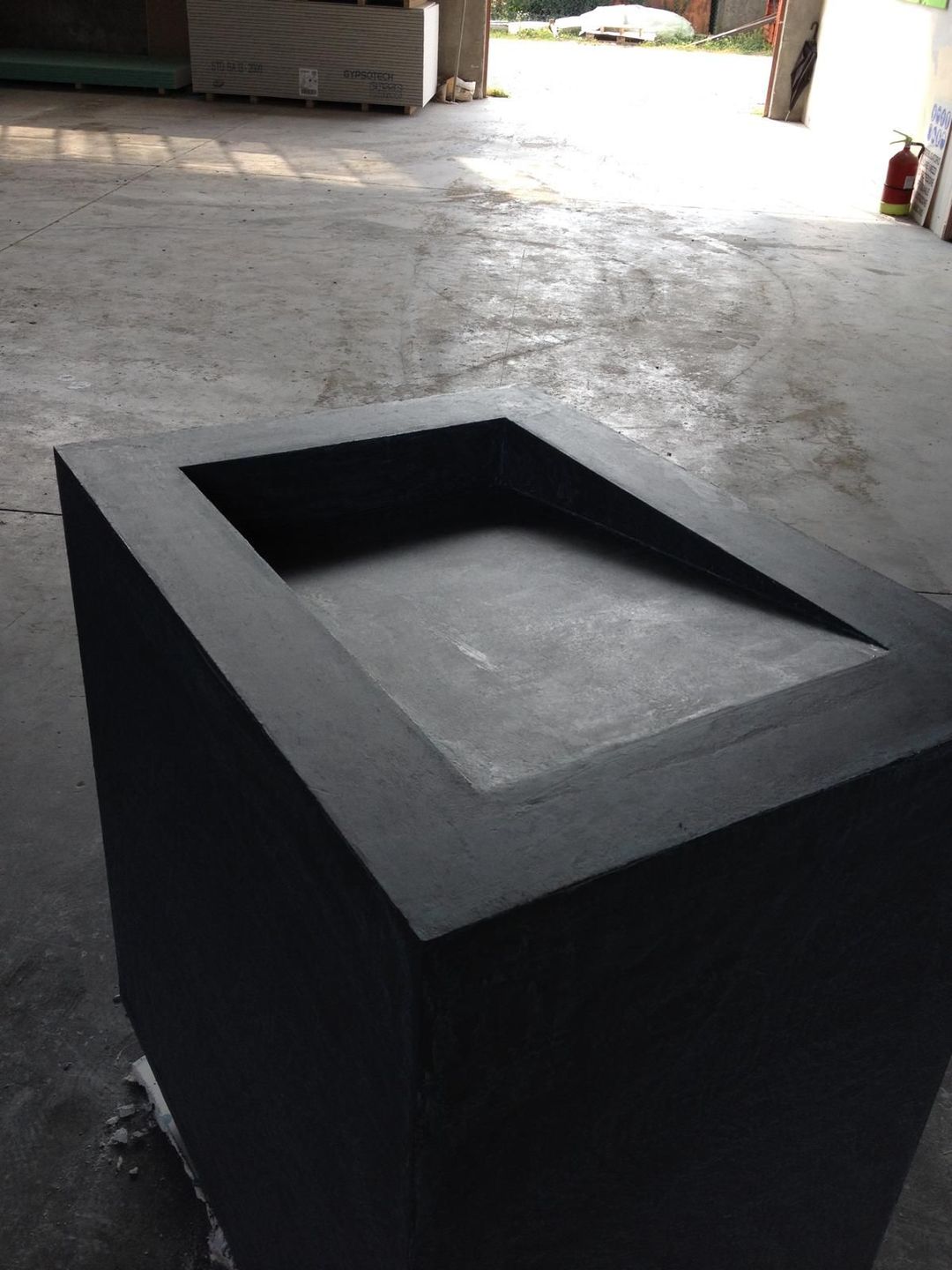 Una vasca o fioriera rettangolare di colore nero con superficie interna inclinata, posizionata in un'officina industriale.