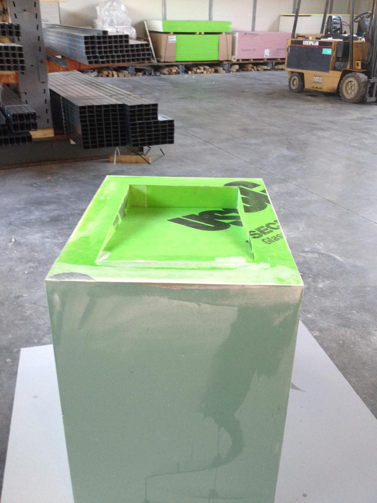 In un ambiente industriale adibito a magazzino, si trova una scatola rettangolare verde con un coperchio incassato, circondata da materiali metallici e un carrello elevatore.