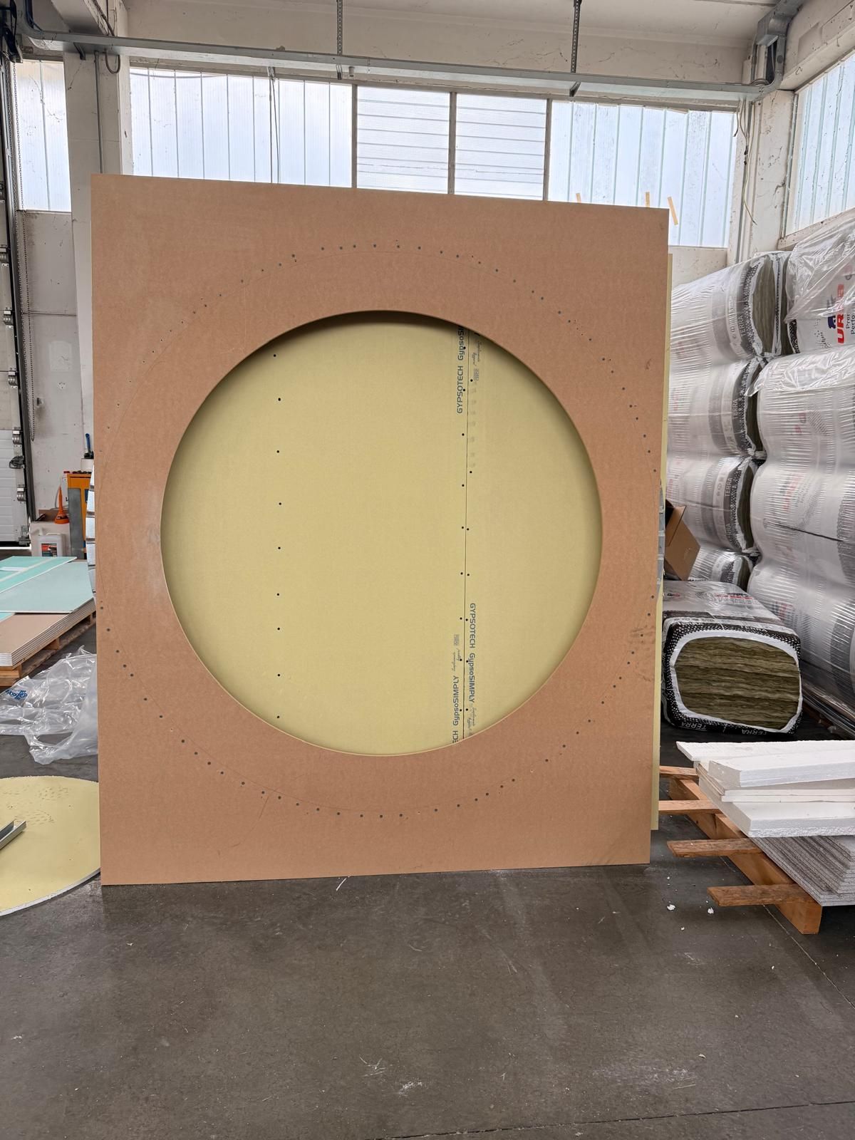 In uno spazio industriale, una grande tavola rettangolare marrone con un ritaglio circolare centrale che lascia intravedere una parete giallo pallido.