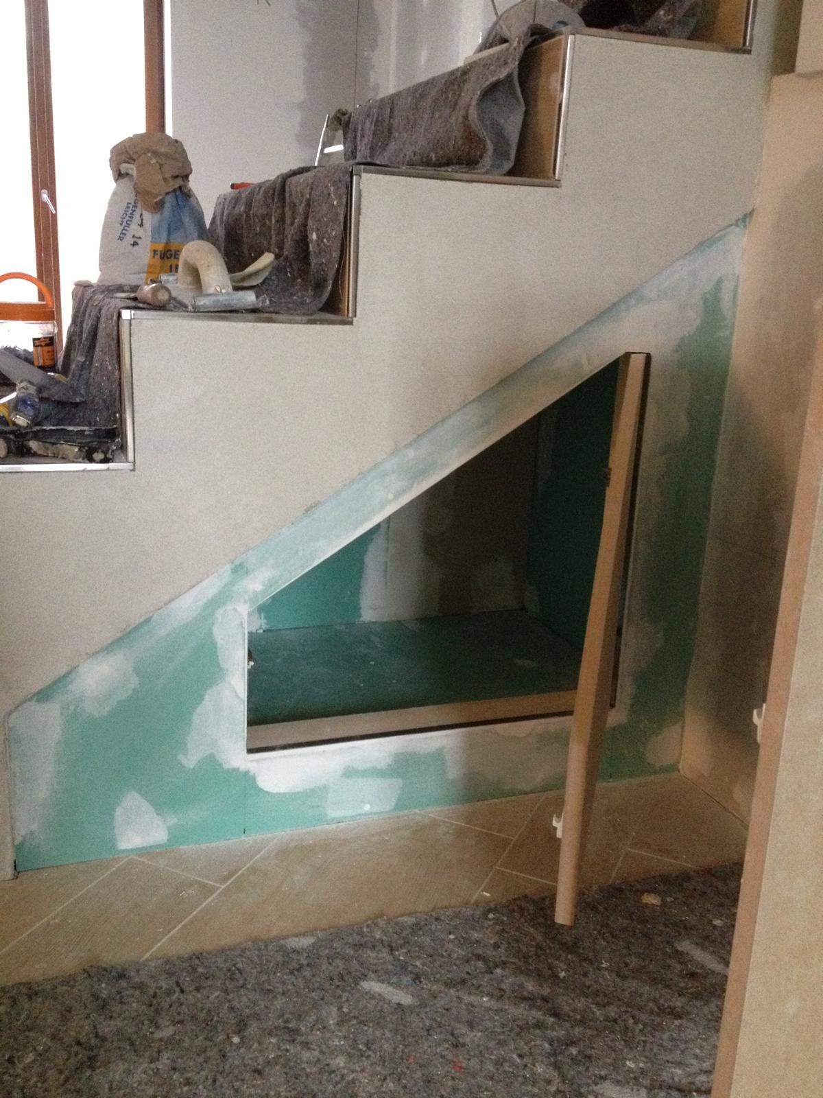 Una scala in costruzione con una nicchia a muro vuota adibita a ripostiglio, incorniciata da cartongesso verde chiaro e profili metallici.