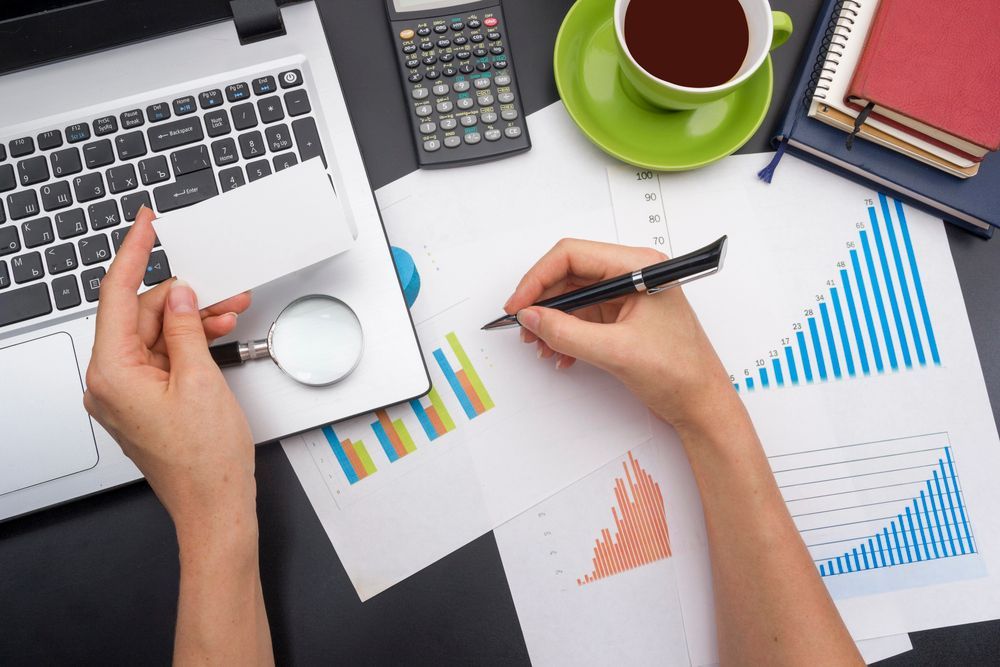 Manos analizando gráficos, computadora portátil, calculadora, café y tarjeta en un escritorio.