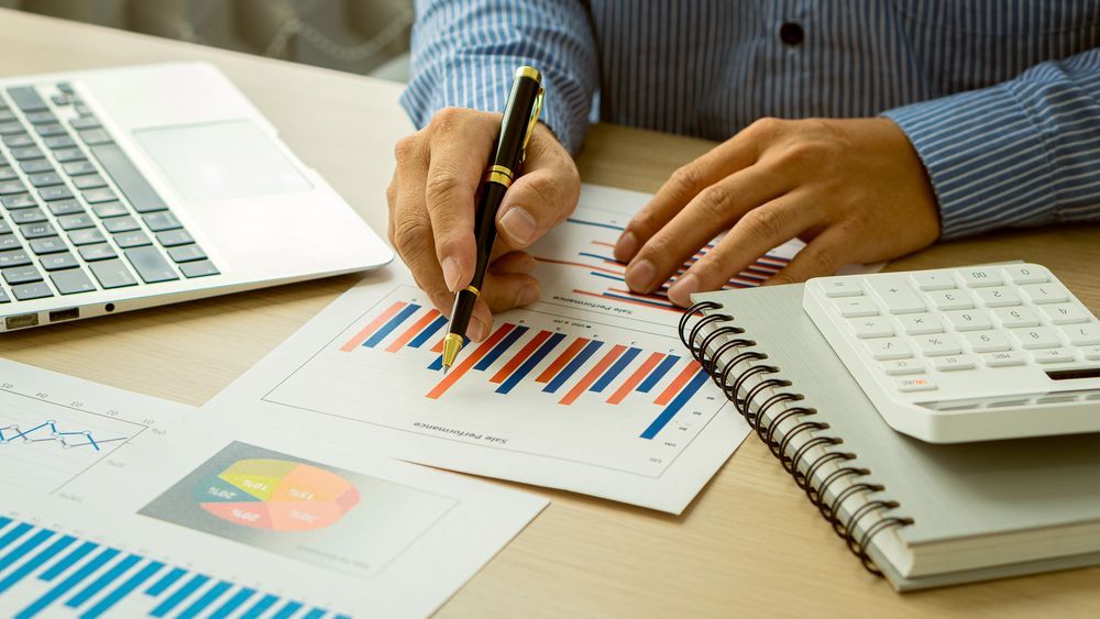 Persona revisando gráficos financieros, usando un bolígrafo, con una computadora portátil, calculadora y cuaderno en el escritorio.