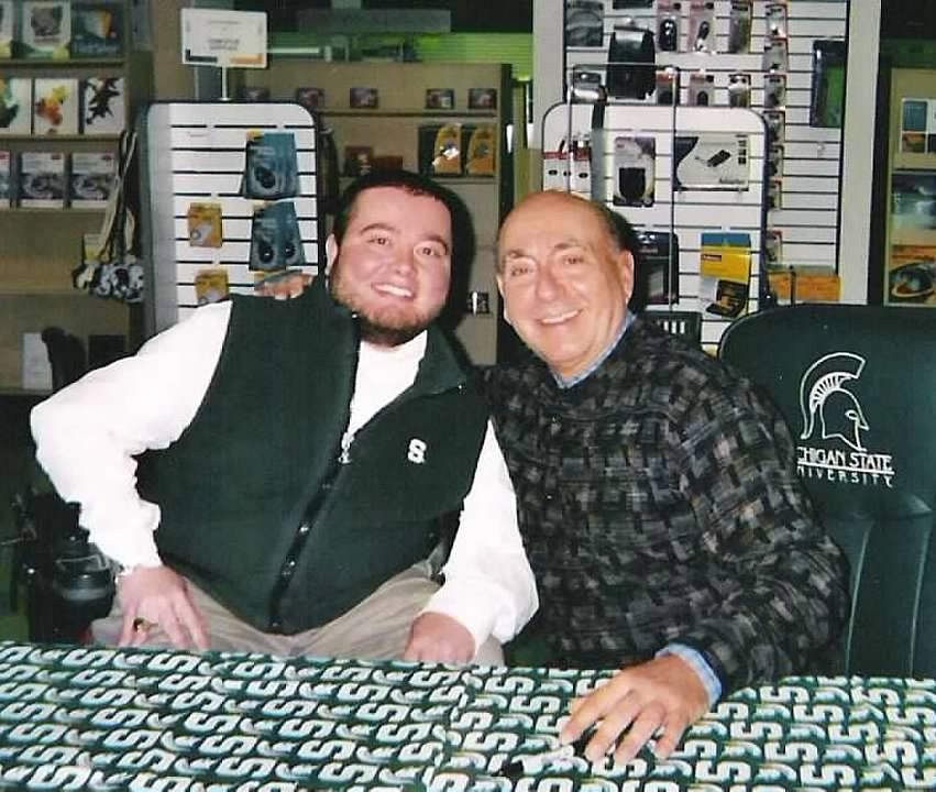 Two men smiling, posing behind a table with a Michigan State University logo. Store shelves in the background.