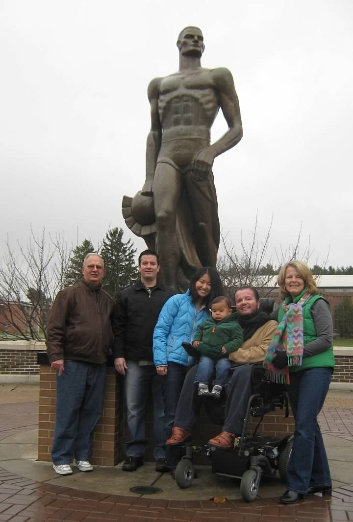 Group poses with The Spartan statue; person in wheelchair in front. Cloudy sky, brick path.