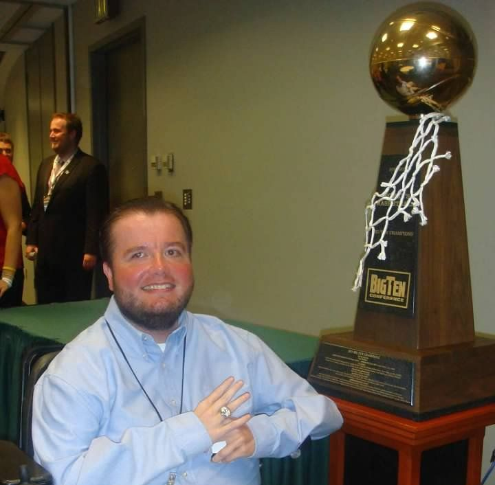 Man smiles beside a Big Ten trophy; gold ball, net, wood base; green table in a hall.