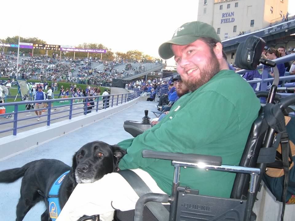 Man in wheelchair with black dog at a football game. Stadium in the background; green and white colors.