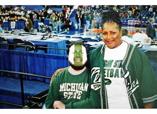 Two people wearing Michigan State gear at a basketball game; one has face paint.