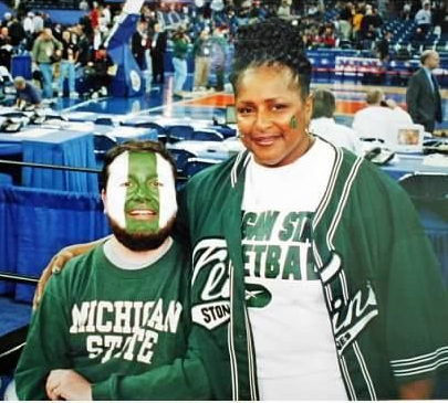 Two people wearing Michigan State gear at a basketball game; one has face paint.