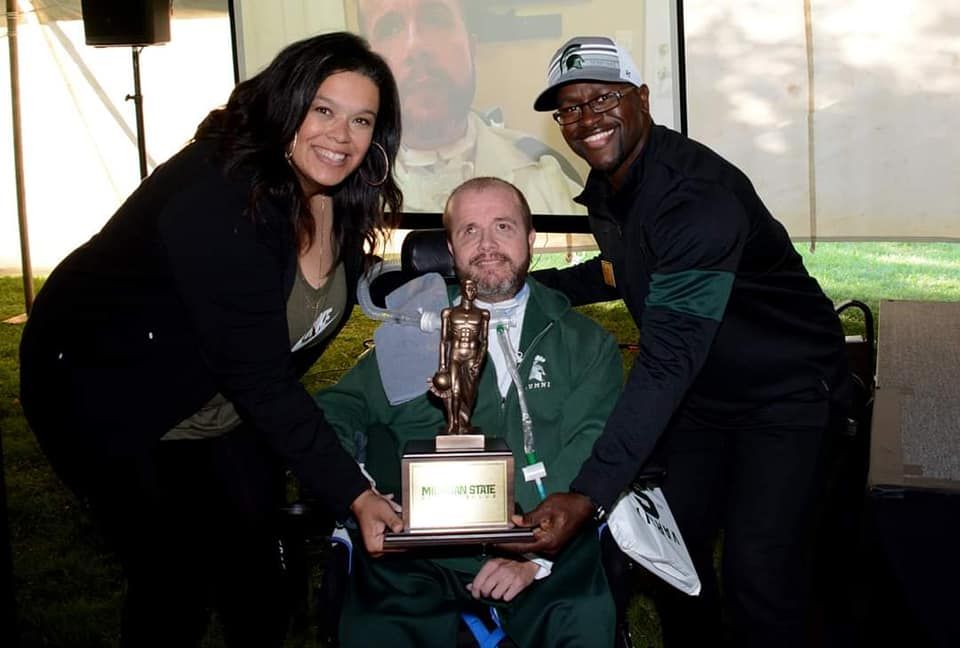 Three people holding an award outdoors; man in wheelchair smiles, flanked by two others, one with a cap.