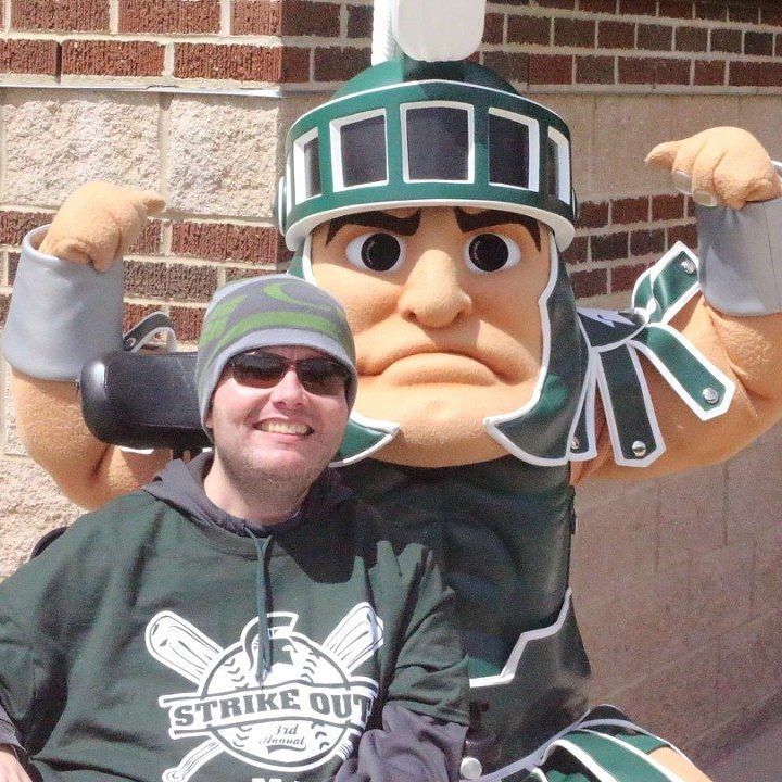 Man in green hoodie smiles with Spartan mascot in front of brick wall.