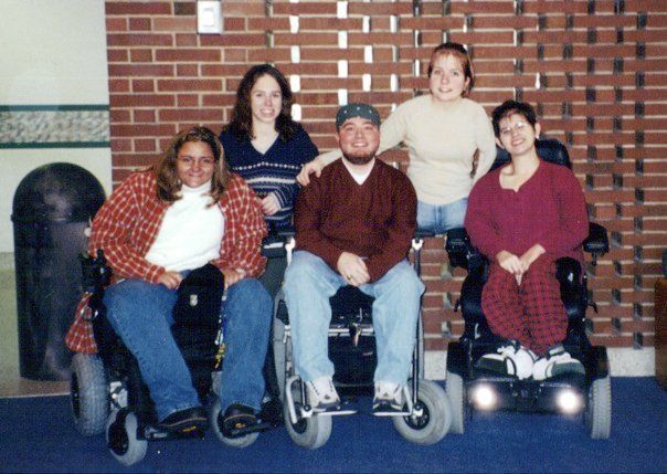 Five people pose together, three in wheelchairs and two standing, in front of a brick wall.