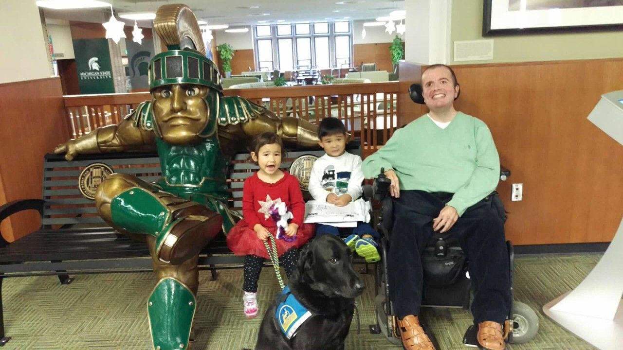 Person in wheelchair with two children and a service dog, posing next to a bronze Spartan statue.
